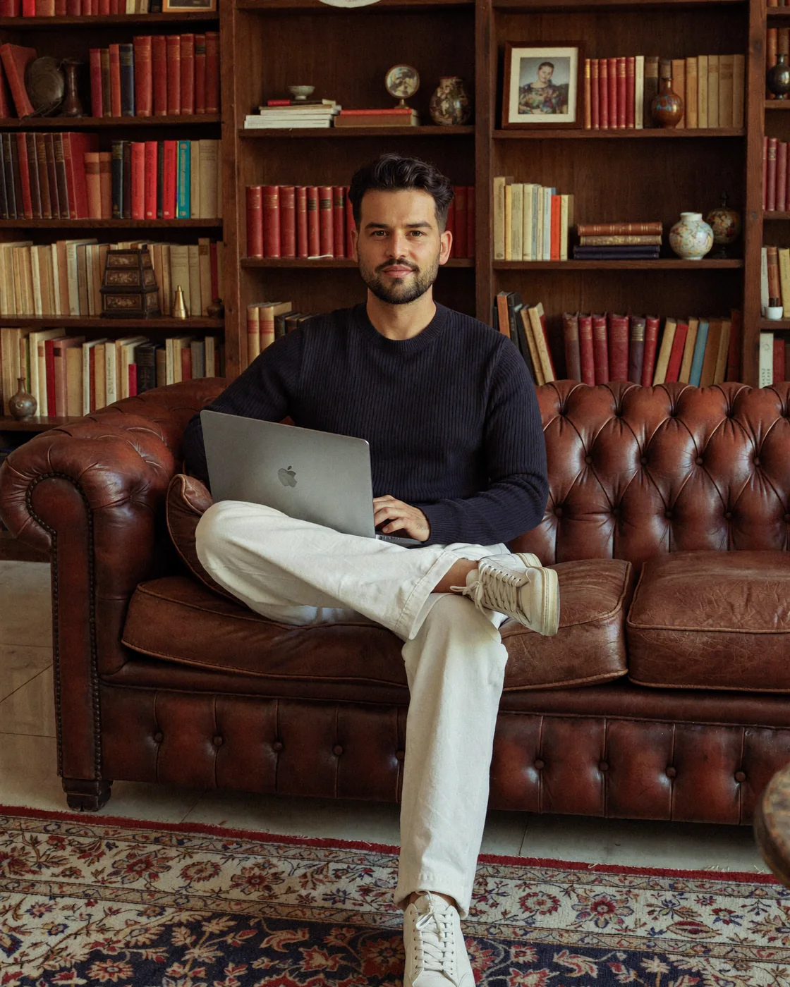 Marco Pöppel sitzt mit Laptop auf einem Ledersofa vor einem Bücherregal